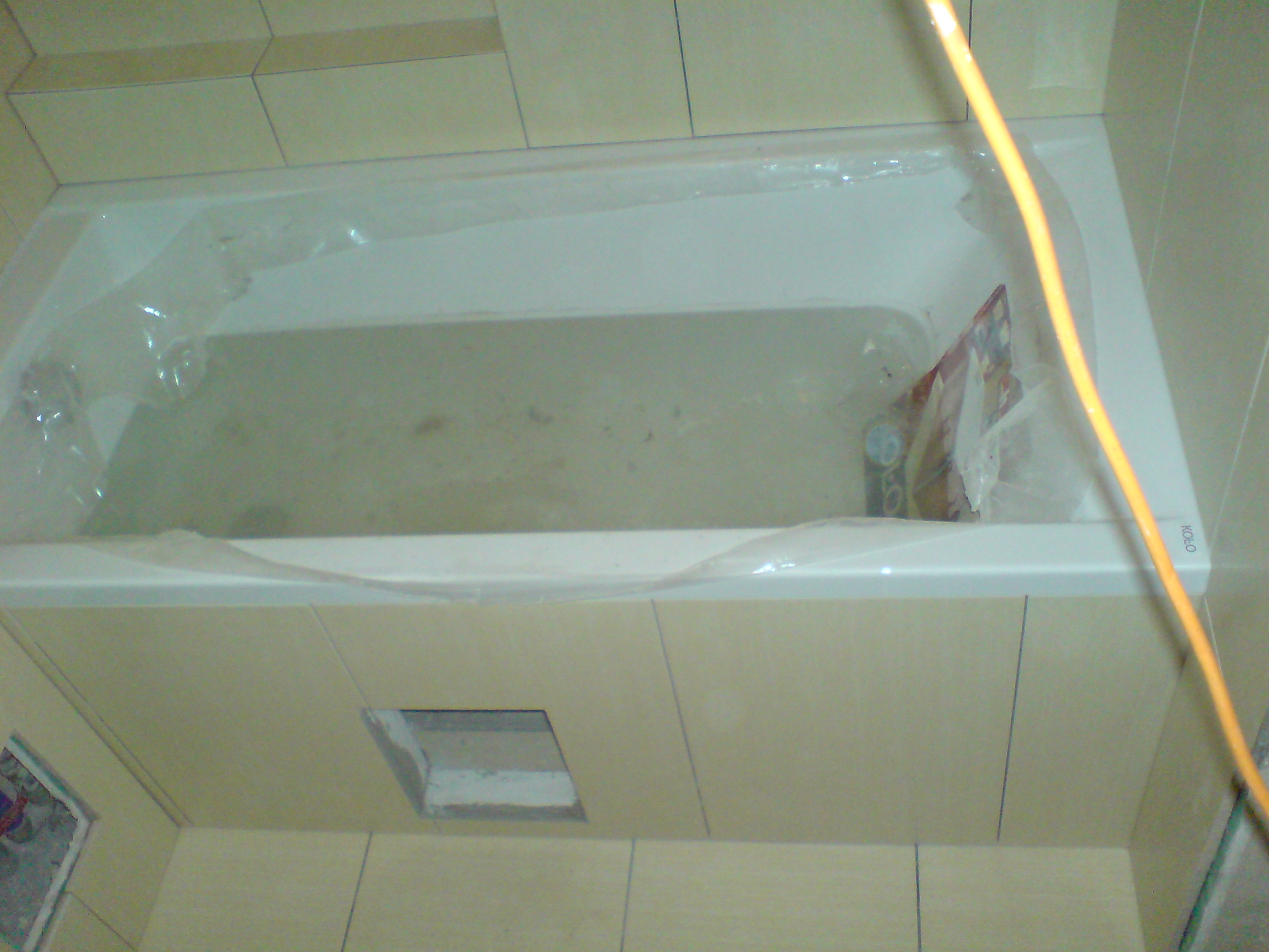 Wanna z obudową w trakcie montażu, zabezpieczona folią, widoczny otwór rewizyjny w obudowie, płytki beżowe na ścianach.